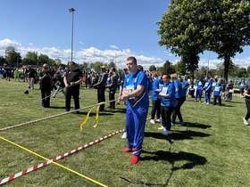 Uczestnicy zawodów sportowych na boisku biorą udział w przeciąganiu liny. Na pierwszym planie mężczyzna w niebieskim stroju sportowym i czerwonych butach trzyma linę. W tle zawodnicy, kibice i sędziowie pod słonecznym niebem.
