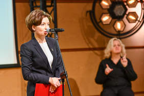 Pokaż zdjęcie: Kobieta mówi do mikrofonu, trzyma złożoną teczkę, obok tłumaczka języka migowego. 