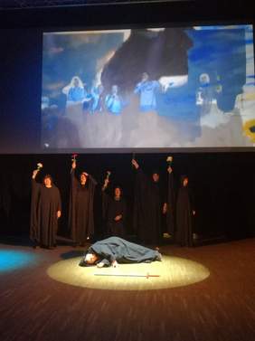 Pokaż zdjęcie: na zdjęciu sala artystyczna na środku stoją osoby ubrane na czarno mają podniesioną  prawą rękę w której trzymają różę jedna osoba leży na scenie  w kapturze przed nią leży rekwizyt w postaci miecza, na telebimie wyświetla się muzyka i  