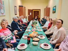 Pokaż zdjęcie: W sali stoi długi stół. Wokół niego siedzą ludzie. Na stole znajdują się różne świąteczne potrawy. 