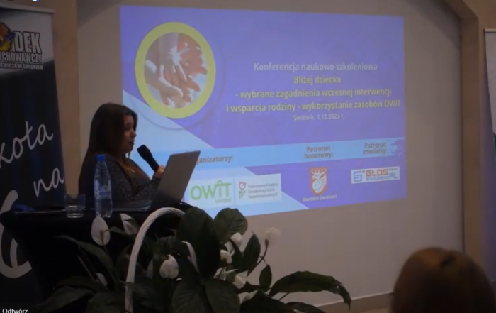 Pokaż zdjęcie: Zdjęcie osoby mającej wykład, kobieta z długimi ciemnymi włosami stoi przy mównicy, w ręku trzyma mikrofon, na ścianie wyświetlana jest prezentacja.