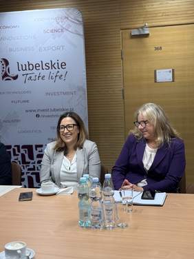 Pokaż zdjęcie: Dwie kobiety siedzą przy stole podczas spotkania. Jedna z nich ubrana jest w jasną marynarkę w kratę, druga w fioletową marynarkę. Na stole znajdują się dokumenty, telefony, filiżanki oraz butelki z wodą. W tle widoczny jest baner promujący województwo lubelskie.