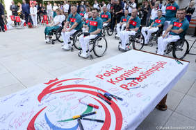 Pokaż zdjęcie: Na stole leży flaga olimpijska z podpisami. W tle nominowani sportowcy, wielu poruszających sie na wózkach.