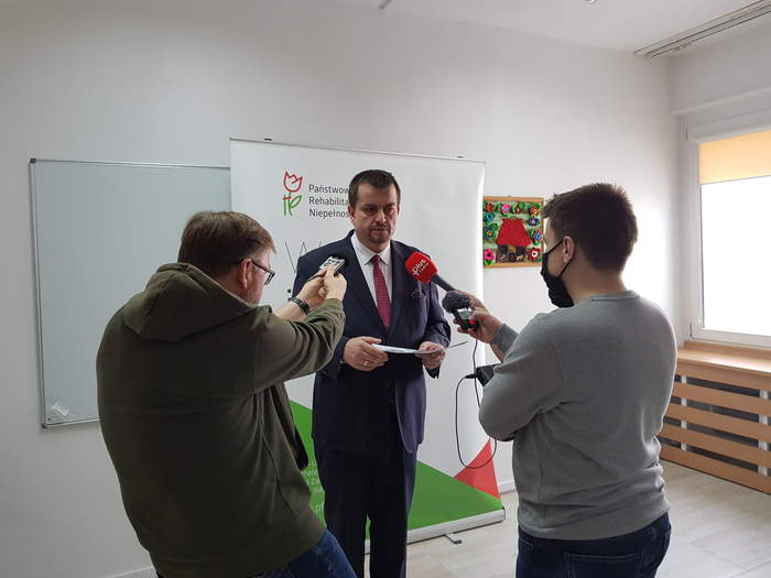 Pokaż zdjęcie: Dyrektor Oddziału Lubuskiego PFRON Andrzej Gonia w trakcie konferencji na stojąco w sali konferencyjnej na tle baneru PFRON, a obok niego stoi dwóch dziennikarzy z mikrofonami