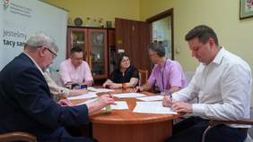 Pokaż zdjęcie: Przy owalnym stole siedzi sześć osób: dwie kobiety i czterech mężczyzn. Na stole leży dużo dokumentów i pieczątek. Osoby rozmawiają ze sobą i podpisują umowy. 