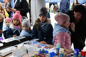 Pokaż zdjęcie: Pod namiotem znajduje się stoisko, gdzie przedszkolaki mają malowane twarze. Na zdjęciu kobieta maluje buzię dziecka.