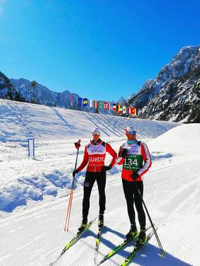 Dwóch narciarzy stoi na trasie biegów, na tle gór, słoneczny dzień
