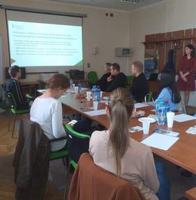 Pokaż zdjęcie: Przy stole siedzą 3 kobiety i 4 mężczyźni spoglądają na ekran na którym wyświetlane są informacje dotyczące spotkania. Po prawej stronie stoi kobieta i przekazuje informacje ze slajdów wyświetlanych na ekranie.