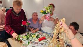 Pokaż zdjęcie: Przy stole siedzą i stoją kobiety, które układają bukiety z kwiatów. Na stole leżą gazety, sztuczne kwiaty i dekoracje. 