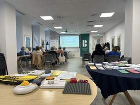 Pokaż zdjęcie: W sali konferencyjnej są ustawione rzędy krzeseł. Siedzą na nich ludzie. W tle widać prowadzącą spotkanie oraz tablicę multimedialną. 