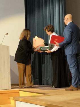 Pokaż zdjęcie: Na scenie kobieta w czarnej marynarce przekazuje grawerton i kwiaty kobiecie w białej bluzce. 