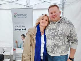 Pokaż zdjęcie: Kobieta i mężczyzna pozują do zdjęcia. Uśmiechają się. Za nimi widać baner Państwowego Funduszu Rehabilitacji Osób Niepełnosprawnych. 