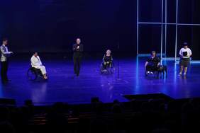 Pokaż zdjęcie: Na scenie stoi mężczyzna ubrany w ciemny garnitur i ciemną koszulę trzyma mikrofon. Obok niego trzy kobiety na wózkach. Dwie ubrane w ciemne sukienki jedna w biały garnitur. Po bokach kobieta i mężczyzna – aktorzy ze sztuki Hairspray.