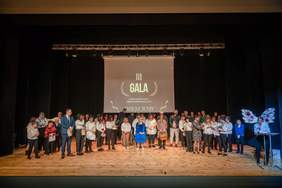 Pokaż zdjęcie: Na scenie Miejskiego Domu Kultury w Rawie Mazowieckiej stoją w półkolu ludzie. Wszyscy są elegancko ubrani. Na ścianie wisi wielki plakat z napisem: III Gala z okazji Dnia Osób z Niepełnosprawnościami. 