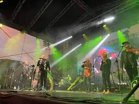 Pokaż zdjęcie: Na dużej scenie stoi dwóch wokalistów z trąbkami oraz chórek składający się z jednej kobiety i dwóch mężczyzn ze skrzypcami. Z tyłu sceny widoczni są instrumentaliści. W tle widoczny jest ekran prezentujący na zbliżeniu głównych wokalistów.