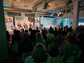 Pokaż zdjęcie: W dużej sali siedzi grupa kilkudziesięciu osób, która przysłuchuje się wypowiedziom uczestników panelu dyskusyjnego. W tle widoczne są banery promujące konferencję, a na dużym ekranie wyświetlona jest także lista uczestników panelu.