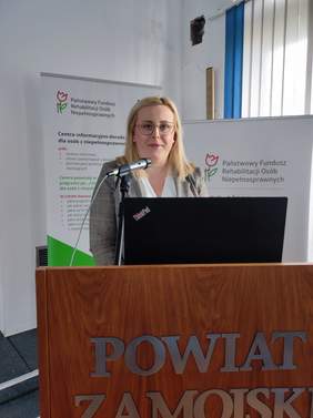 Pokaż zdjęcie: Kobieta blondynka w okularach stoi przy mównicy.