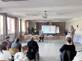 Pokaż zdjęcie: Na zdjęciu widać spotkanie w jasnej sali, gdzie grupa osób uczestniczy w prezentacji na temat dostępności. Kobieta stojąca obok ekranu przedstawia slajd ze zdjęciami barier architektonicznych, a uczestnicy siedzą w półokręgu, słuchając prelegentki.