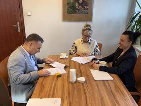 Pokaż zdjęcie: Trzy osoby siedzą przy drewnianym stole w biurze, podpisując dokumenty. Na stole leżą papiery, filiżanki z kawą i pieczątka. W tle widać obraz na ścianie oraz roślinę przy oknie. Atmosfera jest formalna, ale przyjazna.