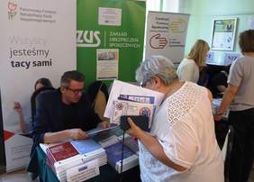 Pokaż zdjęcie: Pracownik Oddziału siedzi przy stoliku i przekazuje stojącemu klientowi broszury oraz informatory. W tle widać nazwy instytucji PFRON, ZUS oraz CPS i NFZ. Obok 4 osoby przy innym stoisku, odwrócone plecami.
