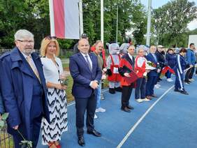 Pokaż zdjęcie: Na stadionie miejskim w Wieruszowie stoją uczestnicy imprezy integracyjnej pod nazwą "Spotkanie przyjaciół". W dłoniach trzymają kwiaty. W tle widać flagę Polski. 