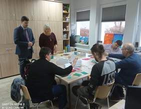 Pokaż zdjęcie: Na stole, przy którym siedzą uczestnicy, znajduje się kilkadziesiąt poskładanych kartek. Uczestnicy zdobią je kolorowym papierem. Wycinają gwiazdki, choinki i świąteczne życzenia. Wysypują brokat udający śnieg.