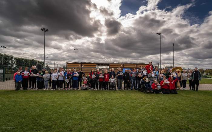 Pokaż zdjęcie: Na pięknej soczystej trawie stoją szczęśliwi uczestnicy i organizatorzy olimpiady. W rękach uczestnicy trzymają dyplomy, mają zawieszone medale. Są wśród nich osoby na wózkach. W tle pochmurne burzowe niebo, przez które przebijają się promie