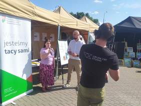 Pokaż zdjęcie: Zdjęcie na festynie, przy stoisku informacyjnym kobieta z długimi włosami odpowiada na pytania redaktora stojącego po jej prawej stronie