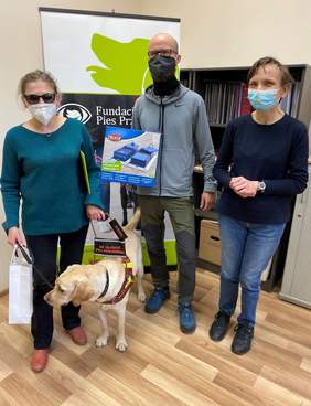 Pokaż zdjęcie: Trzy stojące obok siebie osoby. W środku mężczyzna trzyma w rękach kwadratową planszę z grafiką. Kobieta stojąca z lewej strony trzyma za uprząż psa przewodnika.