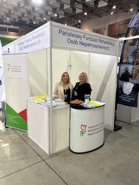 Pokaż zdjęcie: Dwie osoby na stoisku w kolorze białym z kolorowym logiem PFRON. W tle arena sportowa w Szczecinie.