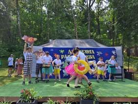 Pokaż zdjęcie: Zielona scena na której trwa występ dzieci. Jako rekwizyty dzieci mają kółka do pływania.