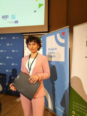 Pokaż zdjęcie: Młoda kobieta z czarnymi kręconymi włosami, ubrana w morelowy garnitur trzyma w ręku czarną teczkę. W tle baner z logo radia Lublin i ZUS.