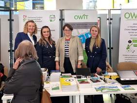 Pokaż zdjęcie: Na tle stoiska informacyjnego pozują cztery kobiety. Na stoliku leżą materiały inforacyjne i ulotki. W tle widać rollupy z Logo PFRON oraz OWiT Kraków. 