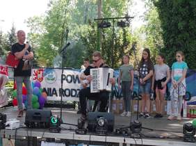 Pokaż zdjęcie: scena w plenerze na środku sceny siedzi chłopak w ciemnych okularach i gra na akordeonie z dala sceny stoją dzieci na scenie  