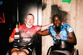 Pokaż zdjęcie: Dwaj bokserzy na wózkach inwalidzkich, trzymający puchary. Są ubrani w koszulki z logo "Seven Stars Paraboxing Night". Jeden z zawodników ma czerwoną koszulkę, a drugi niebieską. Oboje uśmiechają się i obejmują, świętując swoje zwycięstwa.