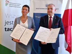 Pokaż zdjęcie: Kobieta i mężczyzna elegancko ubrani stoją w sali konferencyjnej na tle rollupów z logo PFRON oraz PIP. Oboje trzymają otwarte teczki z treścią podpisanego porozumienia.