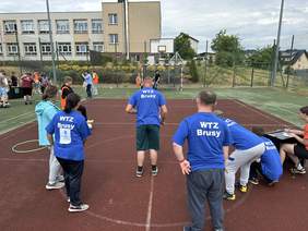 Pokaż zdjęcie: Mężczyzna z w koszulce z napisem WTZ Brusy stoi tyłem do obiektywu i celuje do bramki. Obok stoją inni zawodnicy w tych koszulkach i sędzia.