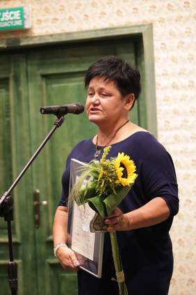 Pokaż zdjęcie: Przed mikrofonem stoi kobieta w kruczo czarnych włosach, ubrana w granatowa sukienkę. W rękach trzyma Oprawiony w ramkę dyplom, statuetkę oraz kwiaty.