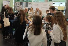 Pokaż zdjęcie: Grupa kilkunastu osób znajduje się przy stoiskach: PFRON i Narodowego Funduszu Zdrowia. Kobieta przy stoisku PFRON udziela informacji zgromadzonym osobom oraz rozdaje ulotki i poradniki. W tle banery PFRON, CIDON, NFZ oraz tablica informacyjna. 