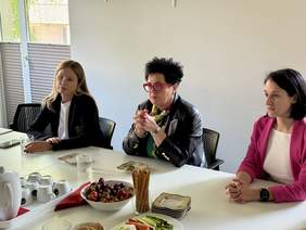 Pokaż zdjęcie: Trzy kobiety siedzą przy stole, patrzą uważnie przed siebie. Przed nimi filiżanki, talerzyki i owoce. Kobieta pośrodku wyróżnia się czarną marynarką i czerwonymi okularami.