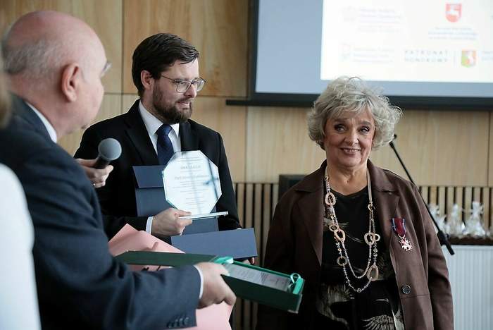 Pokaż zdjęcie: Zdjęcie 3 osób, kobiety i dwóch mężczyzn, mężczyźni wręczają kobiecie uroczyste grawertony