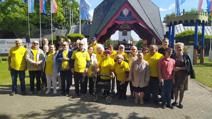 Pokaż zdjęcie: Grupa osób z niepełnosprawnością ubrana w żółte koszulki stoi do zdjęcia na tle budynku