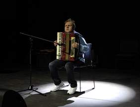 Pokaż zdjęcie: Mężczyzna w błękitnej koszuli i jeansach siedzi na krześle na środku ciemnej sceny, grając na czerwono-białym akordeonie. Artysta jest oświetlony silnym, białym światłem z reflektora punktowego. Przed nim, po lewej stronie, ustawiony jest mikrofon na statywie. Muzyk ma wzrok skierowany lekko w górę, w skupieniu oddając się grze.