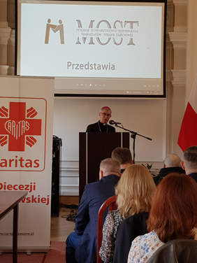 Duchowny, biskup, na mównicy za mikrofonem, nad nim napis MOST