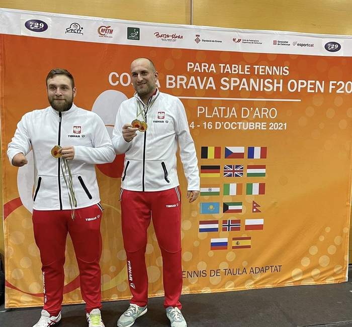Pokaż zdjęcie: Zawodnicy z medalami na tle ścianki promocyjnej Costa Brava Spanish Para Open 2021 w kolorze pomarańczowym