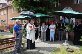 Uczestnicy uroczystości podczas święcenia samochodu stoją na zewnątrz, pod parasolami. Na pierwszym planie stoi ksiądz.