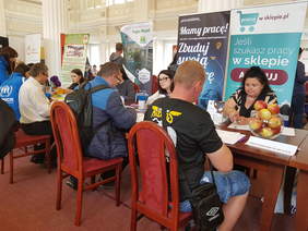 Uczestnicy wydarzenia siedzą po obu stronach długiego stołu, za nimi banery pracodawców