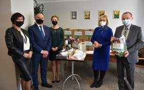 Pokaż zdjęcie: Uczestnicy spotkania: 3 kobiety i 2 mężczyzn, elegancko ubrani, w maseczkach stoją na tle pudełek i paczek z logotypem PFRON. Pośrodku na stole poukładane są produkty, które znajdują się w paczkach