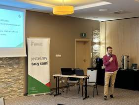 Pokaż zdjęcie: Sala konferencyjna. Po lewej stronie wisi ekran do rzutnika z prezentacją z napisem SOW. Obok baner z logo PFRON z napisem wszyscy jesteśmy tacy sami. Obok stolik z dwoma laptopami. Przy nim stoi mężczyzna w okularach ubrany w koszule i sweter, jasne 