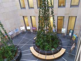 Pokaż zdjęcie: Zdjęcie z góry, w centralnym punkcie duża kompozycja z zielonych kwiatów zwężająca się ku górze, na tle marmurowych ścian stoją sztalugi z wyeksponowanymi pracami plastycznymi. Sztalugi ustawione są w kształt półkola. 